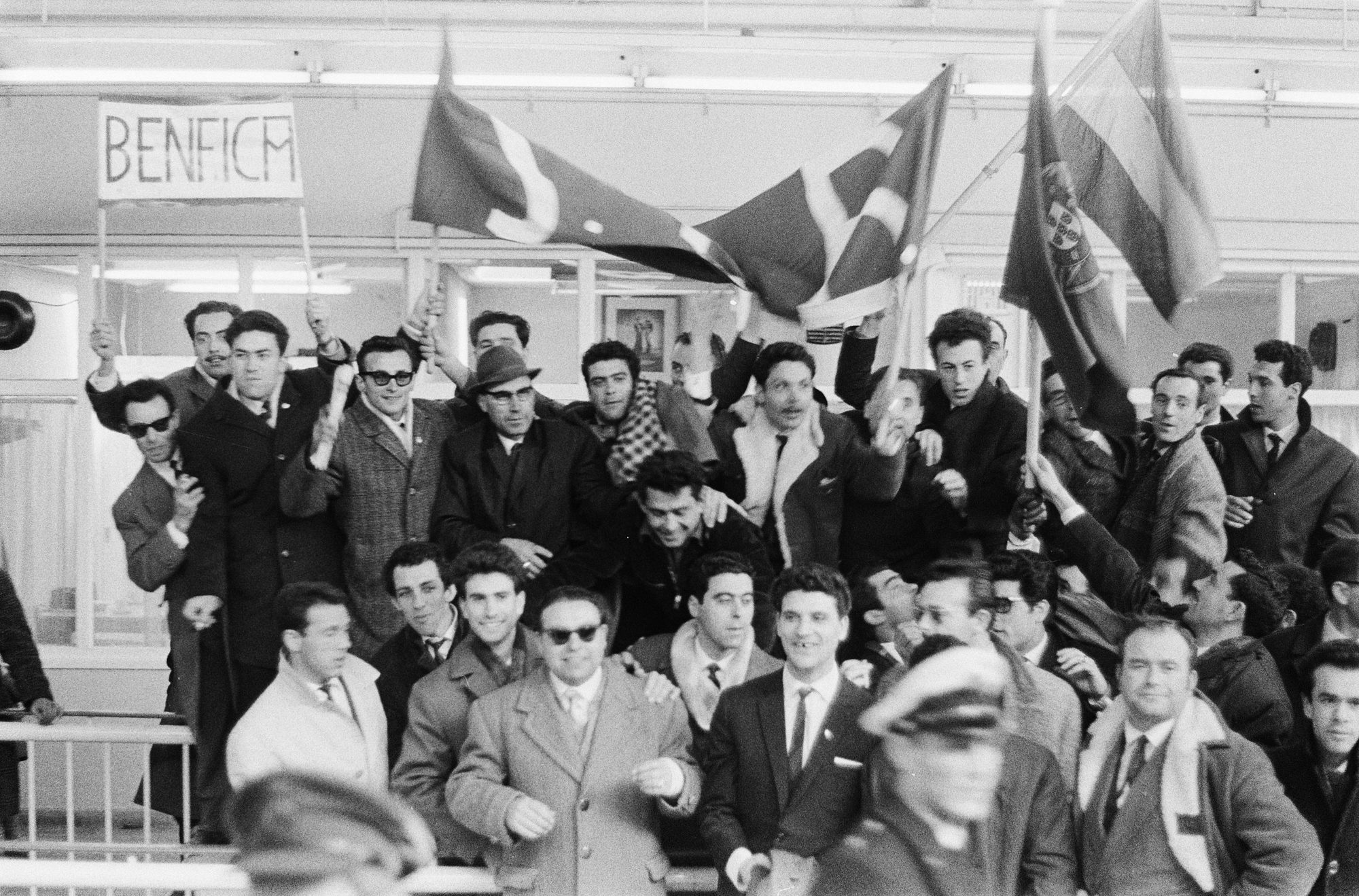 Grupo de homens segurando bandeiras e cartaz escrito ''BENFICA''. Vestem ternos e casacos, com expressões animadas.