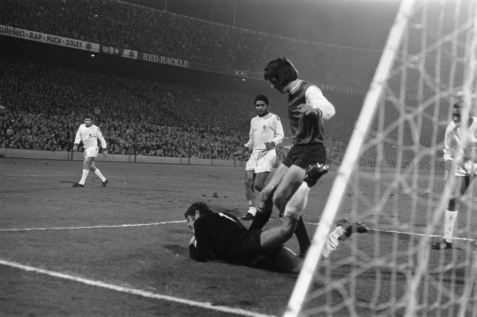 Partida de futebol, jogador de camisa escura próximo ao gol, goleiro no chão segurando a bola, outros jogadores de branco no campo, arquibancadas lotadas ao fundo.
