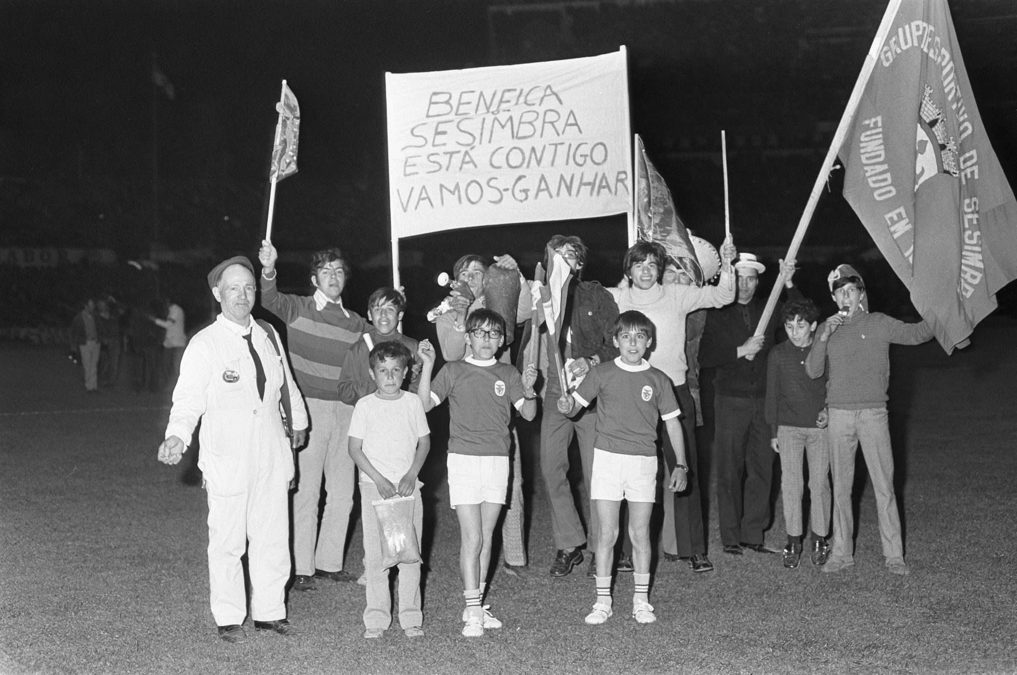 Grupo de pessoas no campo de futebol segurando bandeiras e um cartaz que diz "BENFICA SESIMBRA ESTÁ CONTIGO VAMOS GANHAR". Crianças e adultos estão reunidos em um estádio à noite.