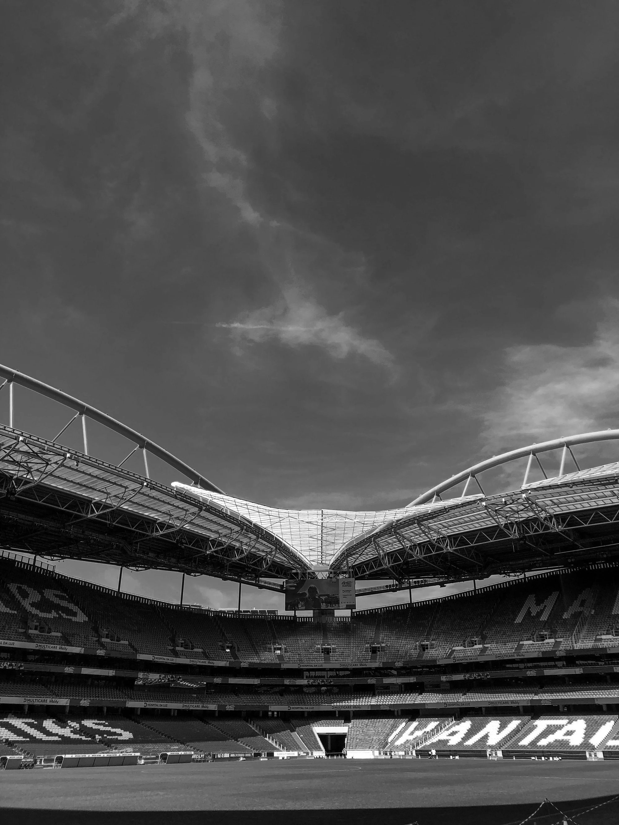 Estádio de futebol moderno com cobertura estruturada, cadeiras vazias e céu parcialmente nublado.