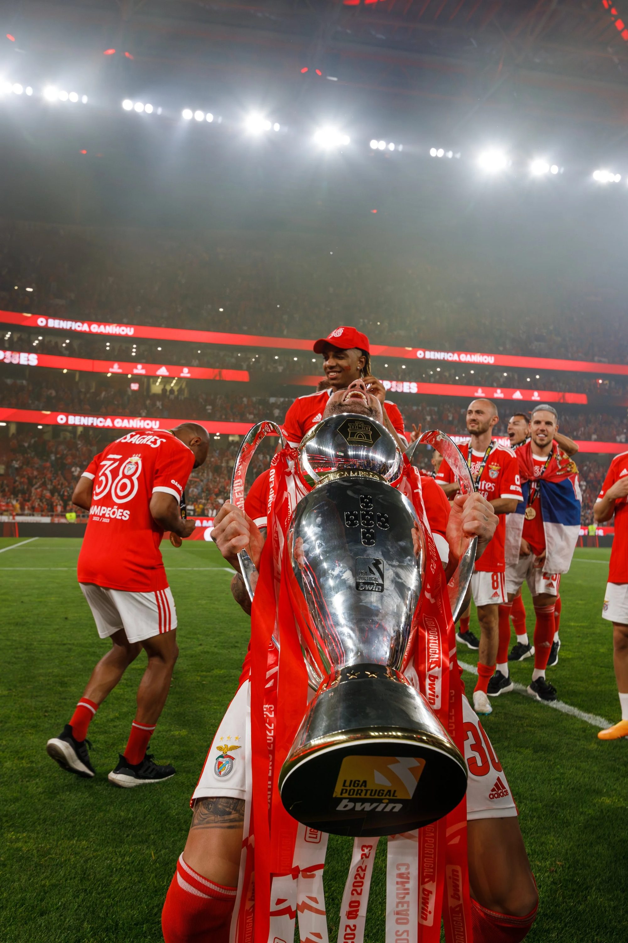 Jogadores de futebol do Benfica comemorando com um grande troféu no estádio, cercados por uma multidão de torcedores. Eles vestem uniformes vermelhos com detalhes em branco.