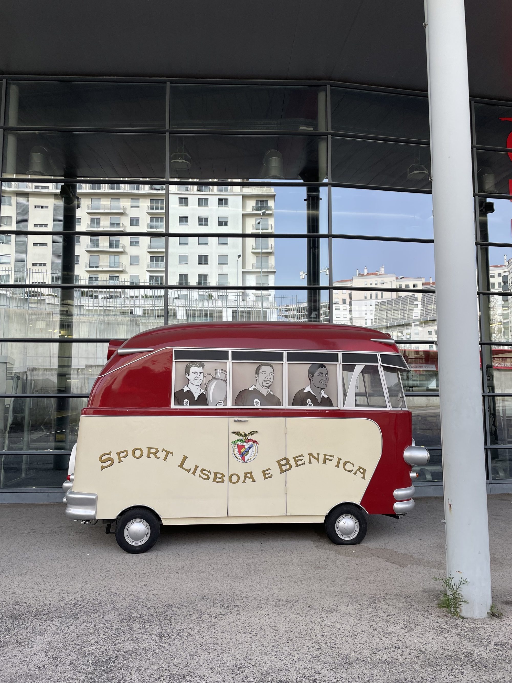 Veículo antigo estilizado com o logotipo e nome "Sport Lisboa e Benfica" em sua lateral, em frente a um prédio moderno com janelas de vidro.